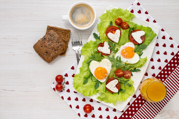 Gala breakfast for two on Valentine's Day. A plate of heart-shaped fried eggs, tomatoes, cheese, coffee and juice on a white wooden table covered with a napkin. View from above. Space for text.