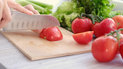 hands using kitchen knife cutting fresh tomato on wooden cutting board, with sprig of tomatoes and lettuce on background