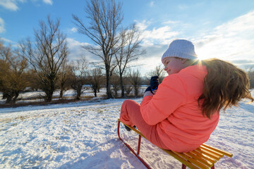 Child girl in an orange ski suit sits on a sled and prepares to go downhill