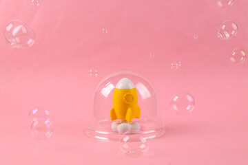 Fun space rocket model under transparent dome and soap bubbles on pink background. Protection,...