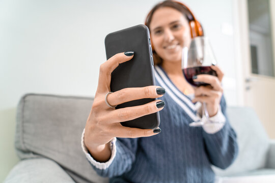 A young woman toasts with friends over a video call with red wine from the sofa at home
