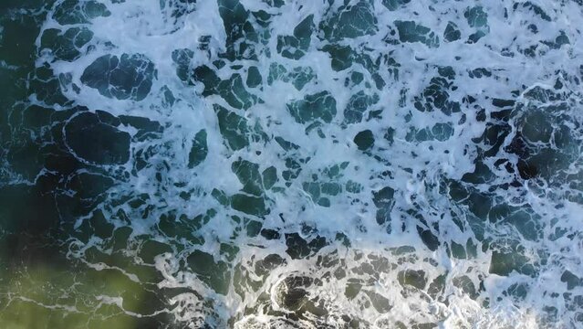 Birds eye view of a beautiful lime green and dark blue ocean as waves crash against waves producing sea foam against dark, slippering rocks as the afternoon sunset casts dark, harsh shadows