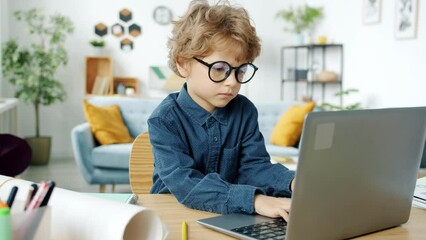 Serious child wearing eyeglasses using laptop typing concentrated on activity in apartment. Modern technology and childhood concept.