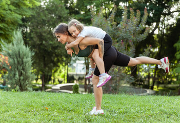 Athletic mom exercising with her little daughter on her back outdoors, healthy lifestyle, fitness...