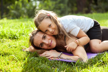 Fit mom with daughter on her back lie on the mat and look at the camera outdoors. Active family.