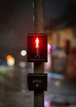 Red man stop sign illuminated at night on traffic light zebra crossing with road and streetlights out of focus behind. Pedestrians waiting for green man to show in rain and dark.