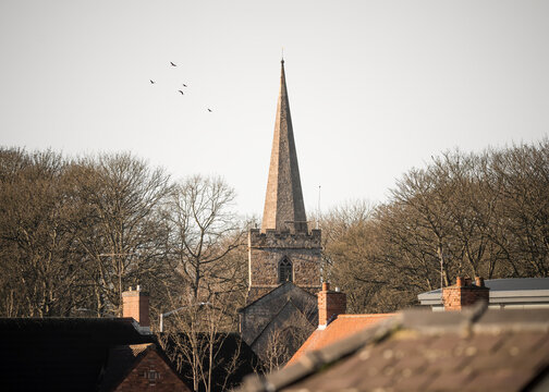 Church stone pointed spire in village centre rising above residential houses of urban town. Sunset and birds in the sky.