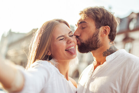 Selfie portrait of a young couple in love. Man kisses his girlfriend on cheek in city. Having fun