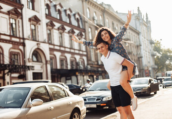 Fototapeta premium Couple in love. Man carrying girl on his back in the street. Smiling man with beautiful young woman, ride piggyback, having fun together. Relationship concept.