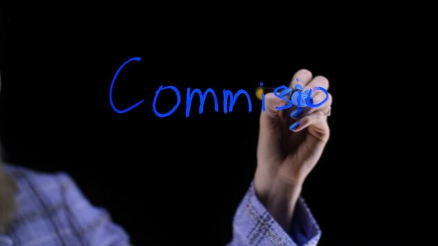 The word commission on a black background. A girl with blonde hair looks at the board and writes the word in blue. Black background near the transparent board.