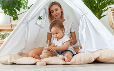 little girl with mamoly plays with a wooden collapsible pyramid for the development of motor skills and thinking while sitting in a wigwam at home © splitov27