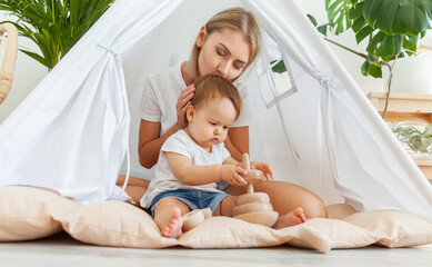 little girl with mamoly plays with a wooden collapsible pyramid for the development of motor skills and thinking while sitting in a wigwam at home © splitov27