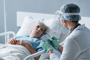 Rear view of young doctor in protective wear writing prescription for senior woman while she lying on bed at the ward