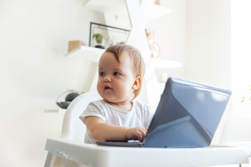 Little funny baby uses a laptop while sitting at the children's table at home