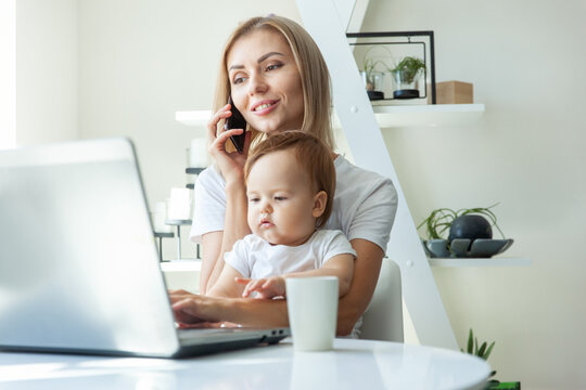 Business Mom With Little Daughter In Her Arms Uses A Laptop And Talking On Phone While Sitting At A Table At Home