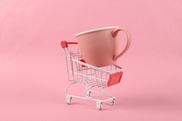 Mini shopping trolley with ceramic cup on pink background