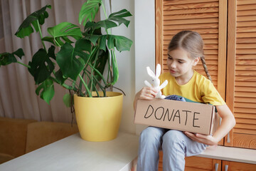 Young girl collect clothes for donate box at home. She holds a toy to add. Second hand and circular...