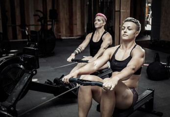 Fototapeta na wymiar Muscular sporty women workout on training simulator in crossfit gym. Sportswomen exercising on rowing machines