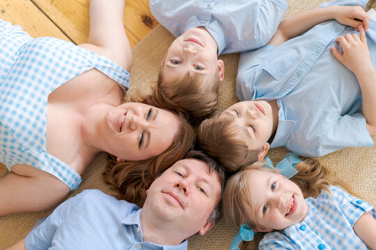 Top view cute children and their beautiful young parents looking at camera and smiling while lying on the floor at home in casual clothes. Big friendly caucasian family on a warm floor looking up