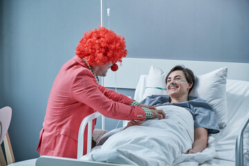 Happy young woman having fun with the clown while lying on hospital bed during her treatment