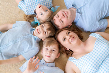 Top view cute children and their beautiful young parents looking at camera and smiling while lying on the floor at home in casual clothes. Big friendly caucasian family on a warm floor looking up