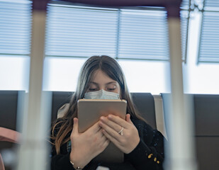 A young blonde Caucasian Girl wearing a mask plays with her tablet at the airport while waiting for her transfer