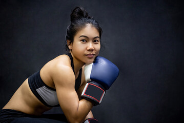 Fototapeta na wymiar Attractive Asian boxer woman sitting and posing with blue boxing gloves in fitness gym. Female boxing class and martial art concept