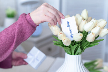 Woman pulling greeting card with Happy birthday words from bouquet of white tulips flowers in a white vase on the table. © flowertiare