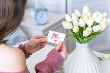 Woman pulling greeting card with Happy mother's day words from bouquet of white tulips flowers in a white vase on the table.