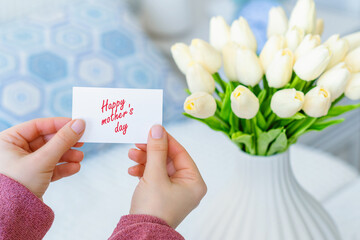 Woman pulling greeting card with Happy mother's day words from bouquet of white tulips flowers in a white vase on the table.