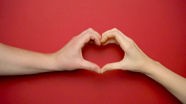 Close-up of two human male and female hands making a heart shaped gesture on a red background. Valentine's Day concept. Romantic gesture of a couple in love.