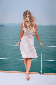 Summer photo from a vacation to the sea from a yacht. Feminine and elegant image with a slim girl with long curly hair turned back in a beautiful plaid summer dress. The observation of the landscape.