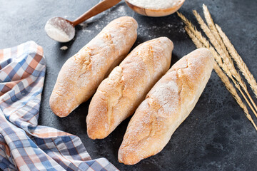 Fresh french baguettes with wheat ears and bowl of flour on dark table, top view