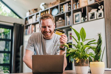 Happy cheerful smiling young adult man doing online shopping or e-shopping satisfied entrepreneur...