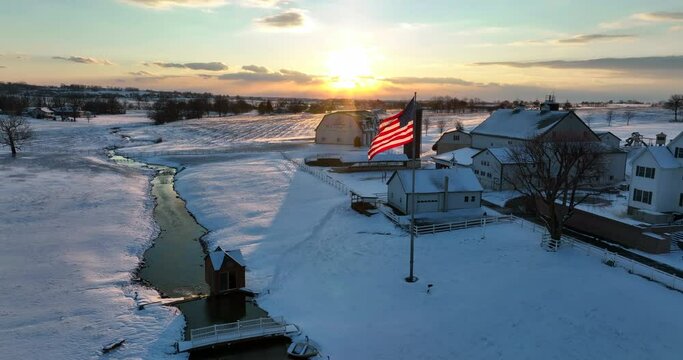 Family farm in winter snow. American flag, creek runs through meadow field at sunset. Beautiful aerial truck shot.