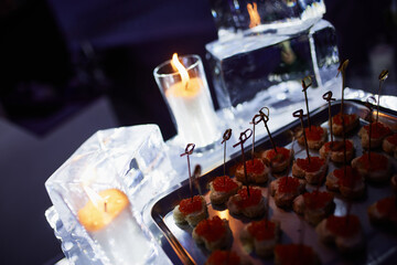 Canape with red caviar on a metal dish on a large block of ice with candles against the backdrop of a festive atmosphere with neon illumination.