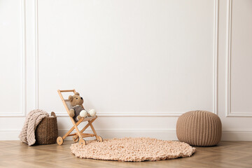 Fototapeta na wymiar Toy stroller with bear, pouf and wicker basket near white wall in child room. Interior design