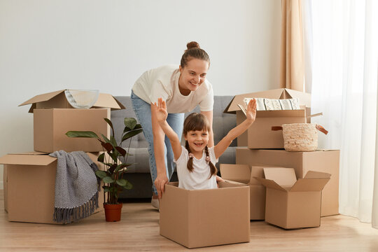 Portrait of happy mom and daughter having fun during moving in a new apartment, expressing optimism, little girl sit in a cardboard box with raised arms, relocation, playing with mother.