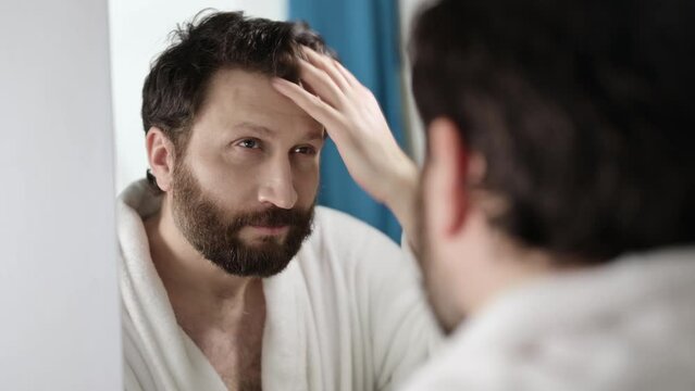 Handsome bearded man combing his hair while looking at himself in mirror after shower. He loves himself, he smiles. A man tunes in to working day, morning rituals.