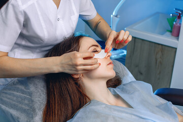 A experienced cosmetologist treats the skin under the client's eyes with collagen patches in a beauty salon.