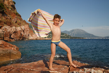 A little boy stands on a stone on the beach with a towel developing like wings in the wind against...