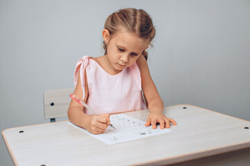 Portrait of a focused attractive child in a dress sitting at a white table and writing numbers in a special sheet of paper. Knowledge concept. photo with noise