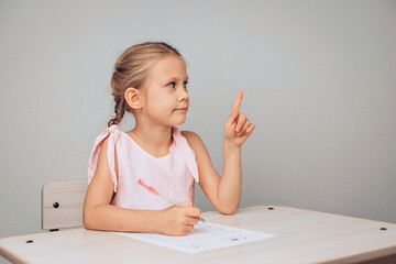 Portrait of a smart pretty girl sitting at a white table drawing and raising her finger up. New idea. Child development. Childhood concept. photo with noise