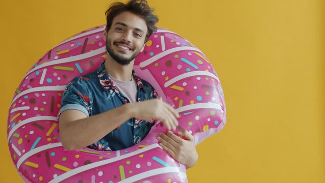 Slow motion of happy Arab guy holding swimming donut and waving hand invition on vacation on yellow color background. Tourism and people concept.