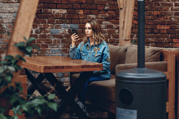Pretty adult woman sitting on a sofa in a cafe against a brick wall background holding a phone and looking into it. Addiction concept