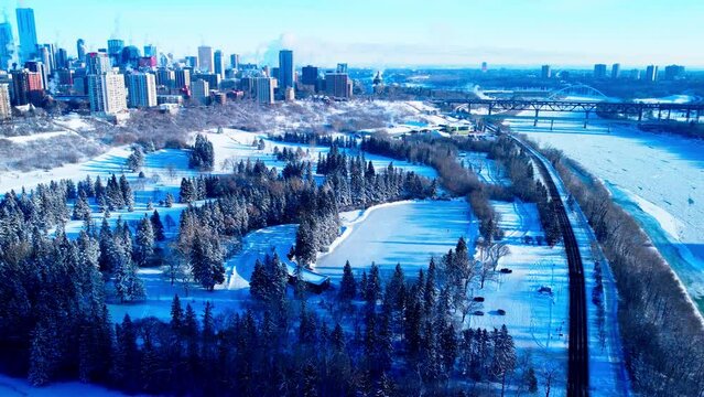 Victoria Park aerial pan out winter snow covered outdoor ice skating oval with only 3 skaters and the Edmonton city skyline in the horizon on a bright sunny day with the shadow of the trees on snow