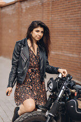 Fototapeta na wymiar Portrait of a sexy force and a bright woman in a black leather jacket and dress posing next to a black retro motorcycle with her knee on the wheel and looking into the distance
