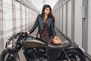 Fototapeta na wymiar Portrait of a bright and daring adult model in a leather jacket and dress posing next to a black motorcycle