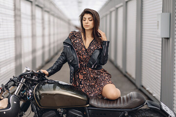 Fototapeta na wymiar Portrait of a sensitive young sweet girl in a black leather jacket and dress posing next to a retro motorcycle with her eyes closed, putting her foot on it against the background of a gray wall