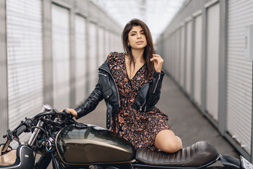 Obraz na płótnie Canvas Portrait of a bright and lovely female model in a leather jacket and dress posing next to a black motorcycle and looking straight into the camera against the backdrop of white walls. Brutality concept